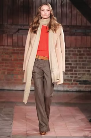 Woman in runway fashion: orange top, tan coat, brown pants, scarf. Brick background.