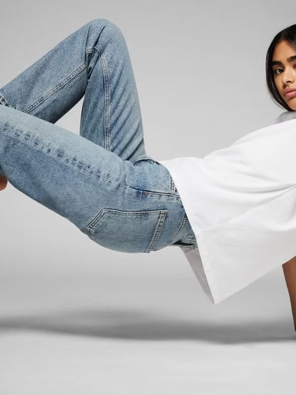 Woman in jeans and white shirt, bent backward with one arm extended, on a white background.