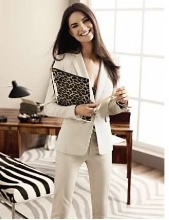 Woman in light suit smiling, holding patterned folder, standing in office.