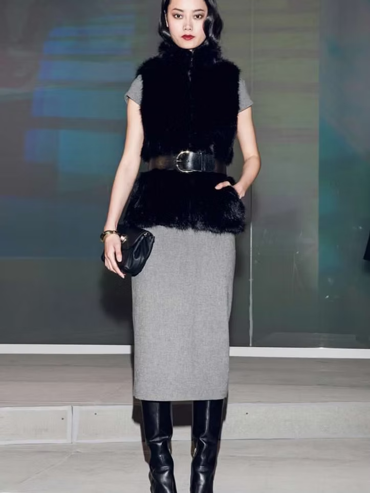 Woman in black fur vest, gray dress, belt, boots, holding a clutch.