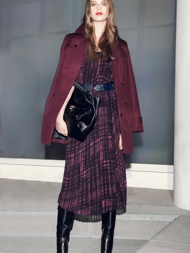 Woman in a maroon coat, plaid dress, and black boots holds a black bag, standing indoors.