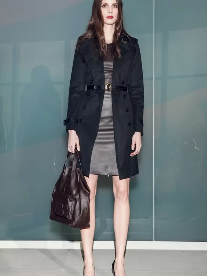 Woman in black coat and dress holding a brown bag, posing in front of a blue wall.