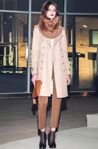 Woman in tan trench coat, brown pants, and fur scarf, holding a clutch, wearing ankle boots. Outdoors.