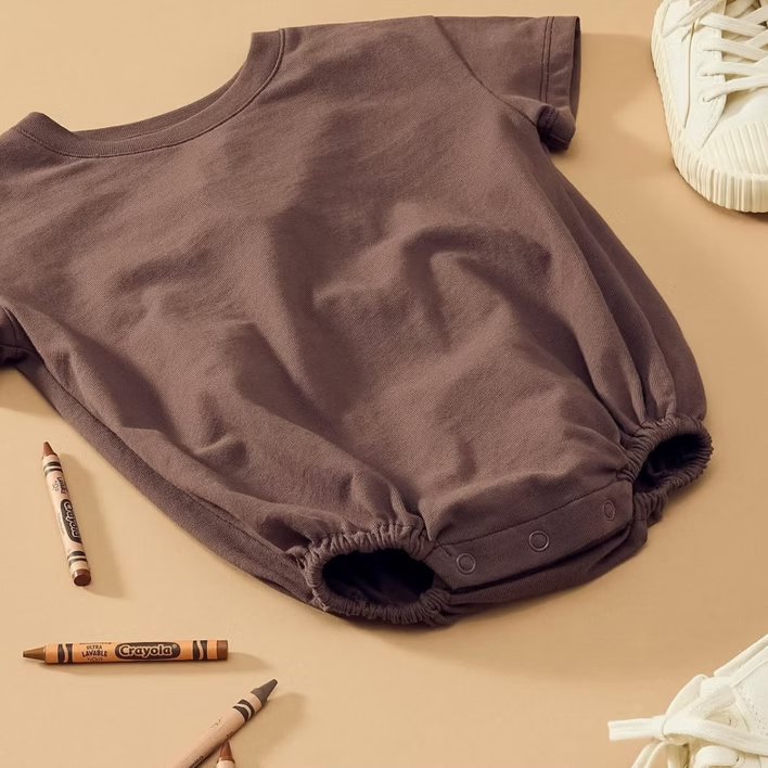 Brown baby romper with crayons and white sneakers on a beige surface.
