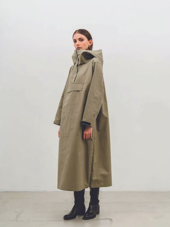 Woman in olive green, hooded, oversized coat and black boots in a studio.