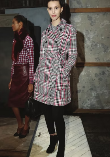 Person in a plaid coat and black leggings posing indoors on a runway-like platform.