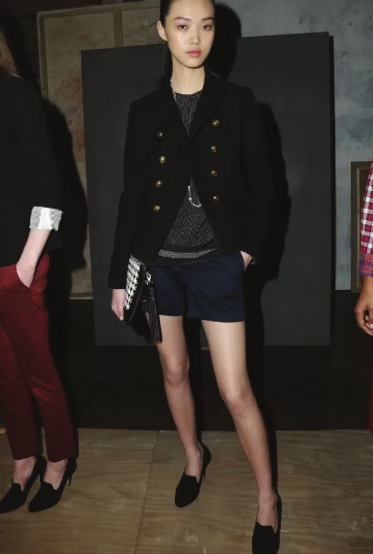 Woman in black coat, shorts, and shoes, holding a clutch, with hands in pockets.