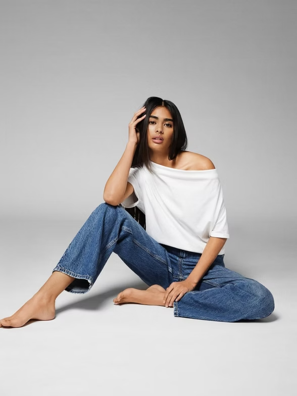 Woman in white off-shoulder top and blue jeans, sitting on a gray floor, hand in hair.