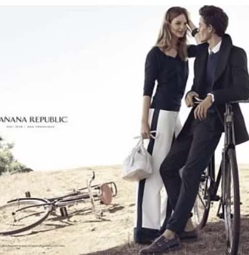 Couple leaning on a bicycle in a field. Woman in black sweater and white pants, man in a black coat.