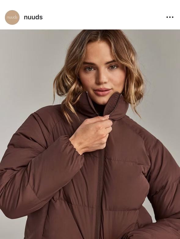 Woman in brown puffer jacket, buttoning up the collar, neutral background.