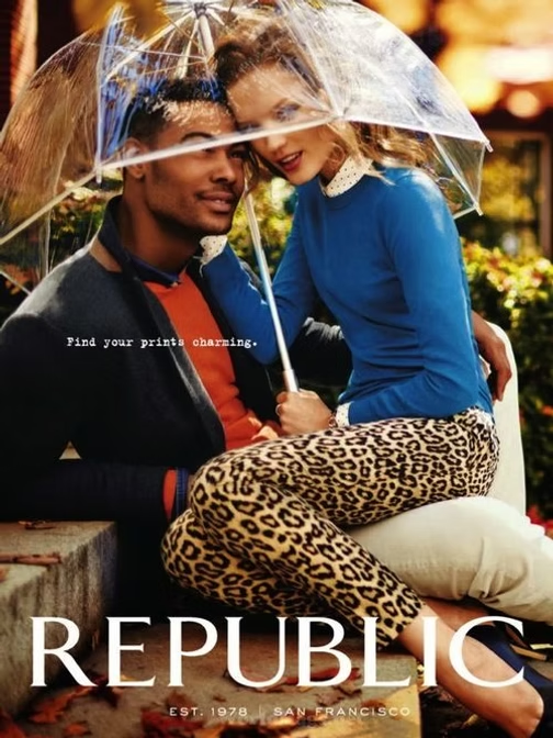 Couple under an umbrella, smiling. Woman in blue sweater and leopard pants, man in jacket. Republic ad.