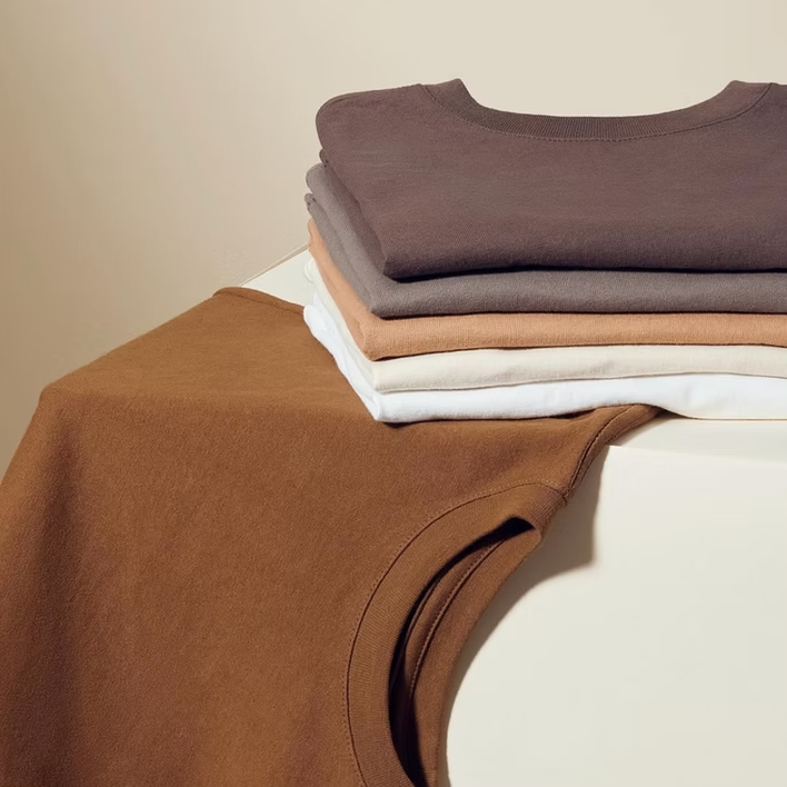 Stacked and folded t-shirts in earth-tone colors on a white surface. One shirt is draped over the side.