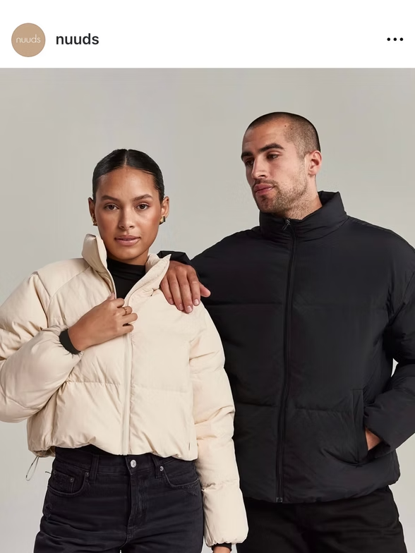 Two people wearing puffer jackets. One is beige, the other is black. Both are standing close together.