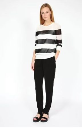 Woman in striped sweater and black pants, hands in pockets, standing against a white backdrop.