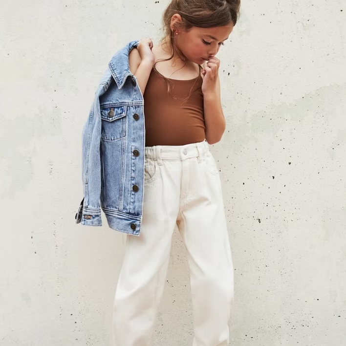Girl wearing brown top, cream pants, and denim jacket. She is leaning against a light wall.
