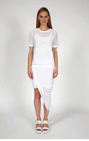 Woman in white outfit: T-shirt, draped skirt, sandals. Standing in front of a white backdrop.