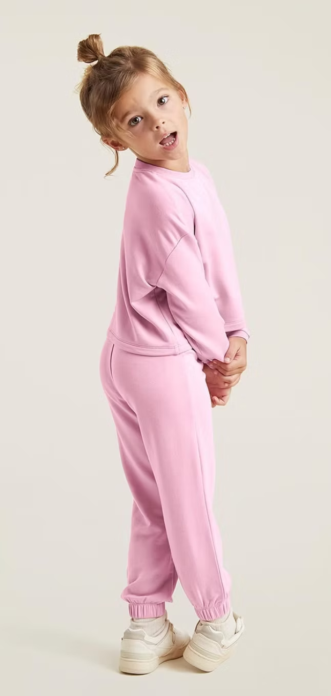 Young girl wearing a pink sweatsuit, smiling, hair in a bun, against a cream background.