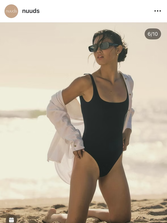 Woman in black swimsuit and sunglasses kneels on a beach, wearing a white shirt.