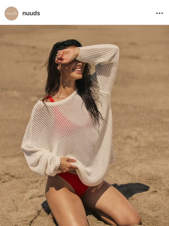 Woman in red swimwear and white sweater shielding eyes on beach, smiling.