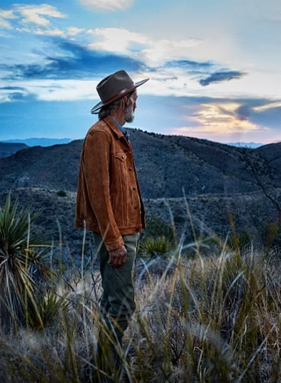 Man in hat and brown jacket looks out over mountain range at sunset.