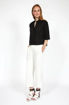 Woman in black top and white pants stands with hands in pockets; white background.