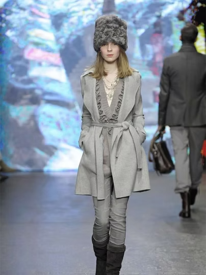 Woman in gray coat, hat, and pants on runway, posing. Another person walks in background.