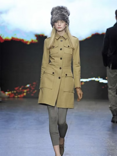 Woman in a tan coat and fur hat on a runway, wearing gray leggings and brown shoes.