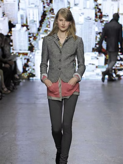 Woman in gray blazer, pink top, and gray pants walks runway in front of cityscape.
