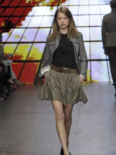 Woman in fashion show; gray jacket, black top, short skirt, leopard belt, runway.
