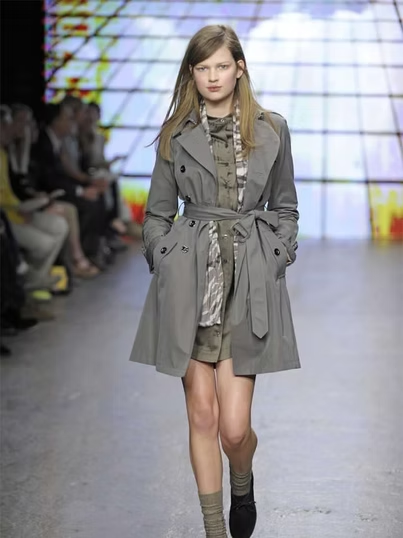 Model in gray trench coat, walking on runway, colorful backdrop.