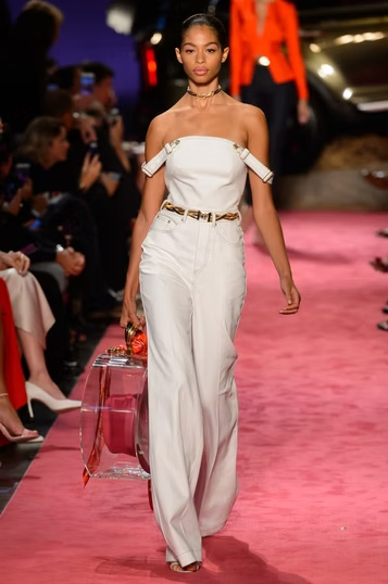 Woman in white jumpsuit, walking runway, pink carpet.
