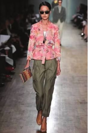Woman on runway in floral jacket, cargo pants, sunglasses, holding clutch, brown boots.