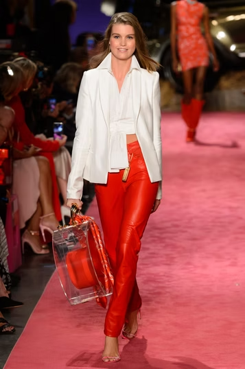Model in white blazer, red pants, and clear bag walks the runway.