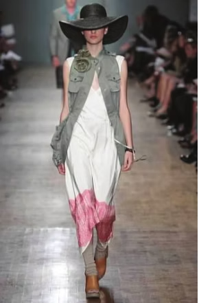 Model on a runway wearing a wide-brimmed hat, long vest, white and pink dress, and boots.