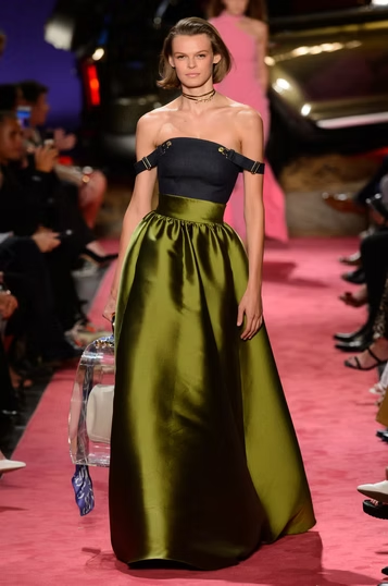 Woman in olive green gown, navy top, walking runway. Pink carpet, fashion show.