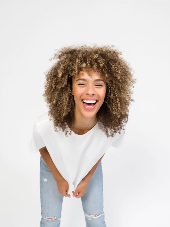 Woman with curly hair smiles, leans forward, wearing white tee and jeans, against white background.