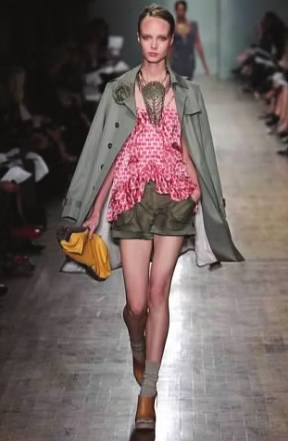 Model in pink top, khaki shorts, olive trench coat, walking runway, yellow bag.