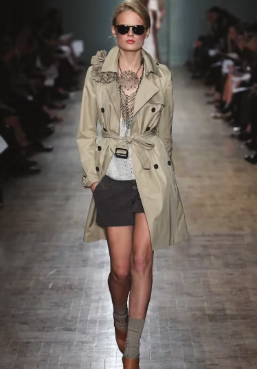 Model in tan trench coat, shorts, and sunglasses walks the runway.