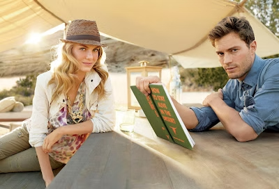 Woman in hat and man reading a book, leaning on a counter outdoors with sunlight.