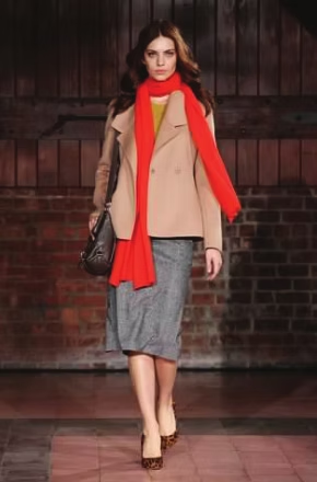 Woman wearing tan jacket, red scarf, gray skirt, and heels, walking in front of a brick wall.