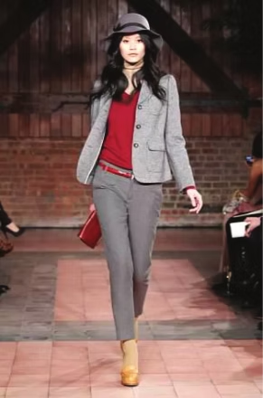 Woman in gray suit, red sweater, hat, and platform shoes walking on a runway.