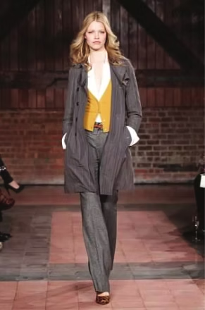 Woman modeling a grey coat over a yellow vest and white shirt with matching pants and brown shoes on a runway.