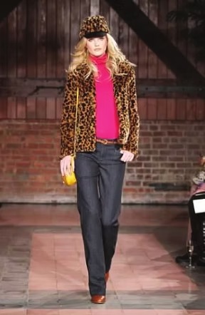 Woman in leopard print jacket, pink turtleneck, and dark jeans walks a runway.