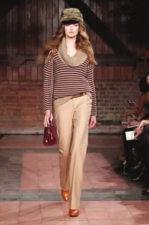 Model wearing a striped sweater, tan pants, and a hat walks a runway with a red brick backdrop.