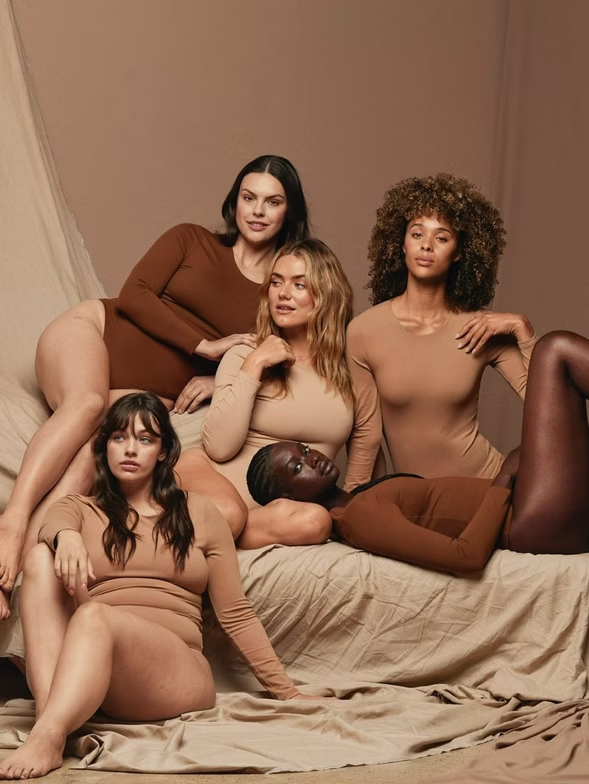 Women in skin-tone bodysuits pose on a bed. Soft lighting, neutral background.