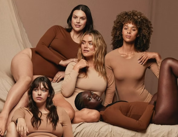 Five people in neutral-toned bodysuits lie on a bed. Various skin tones are represented.
