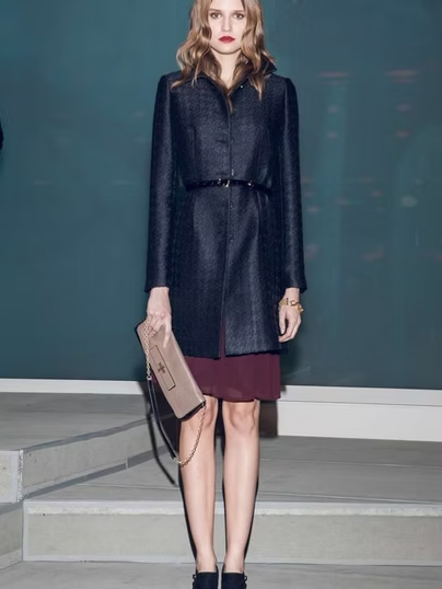 Woman wearing a navy coat with a thin black belt, holding a beige purse, standing on a step.