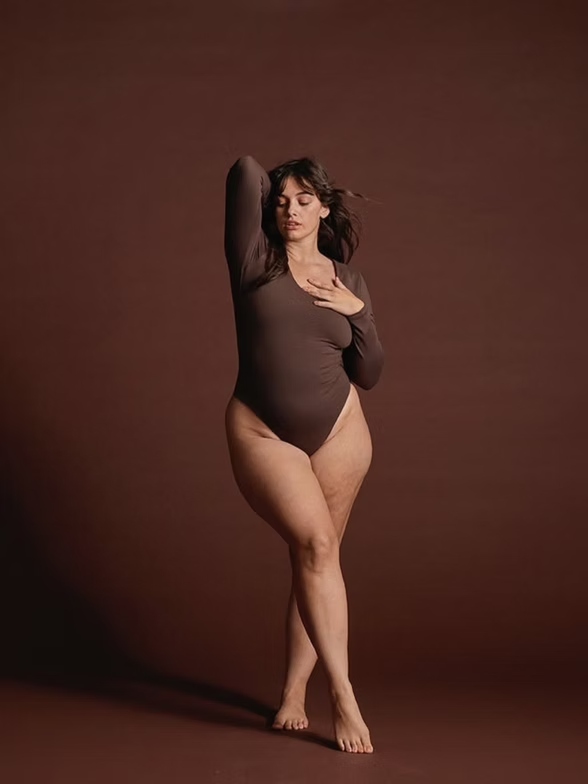 Woman in brown bodysuit, standing with crossed legs, hand on chest, brown background.