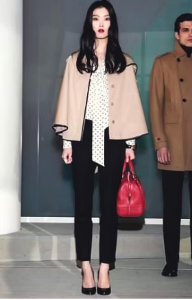 Woman in cape and black pants holding a red bag, standing with a man in a brown coat.