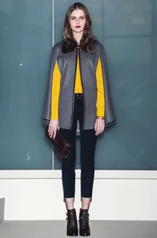 Woman in gray cape and yellow top, holding clutch. Black pants and boots.